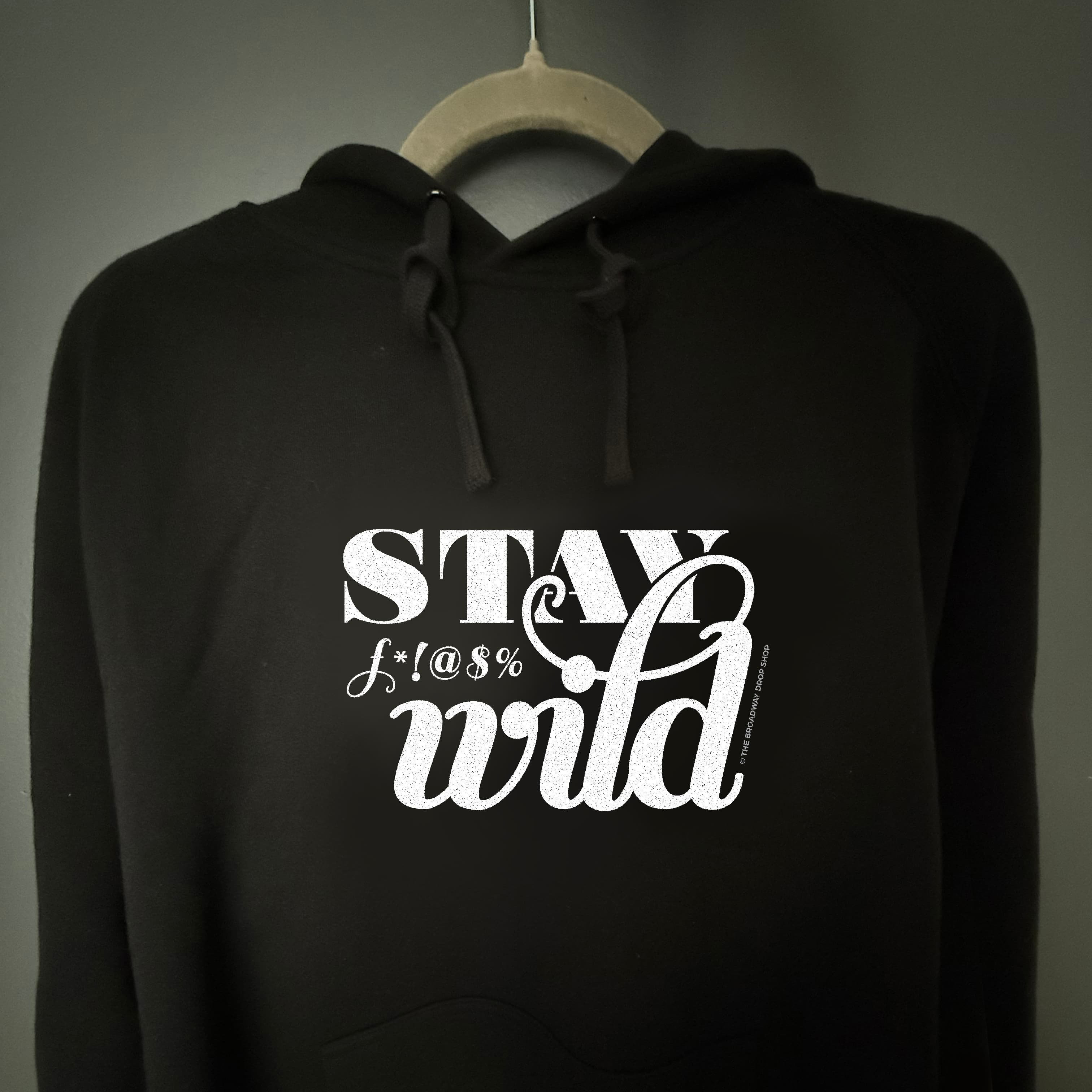 Woman making rock-on gesture wears black hoodie with white "Stay F***'in Wild" text print.