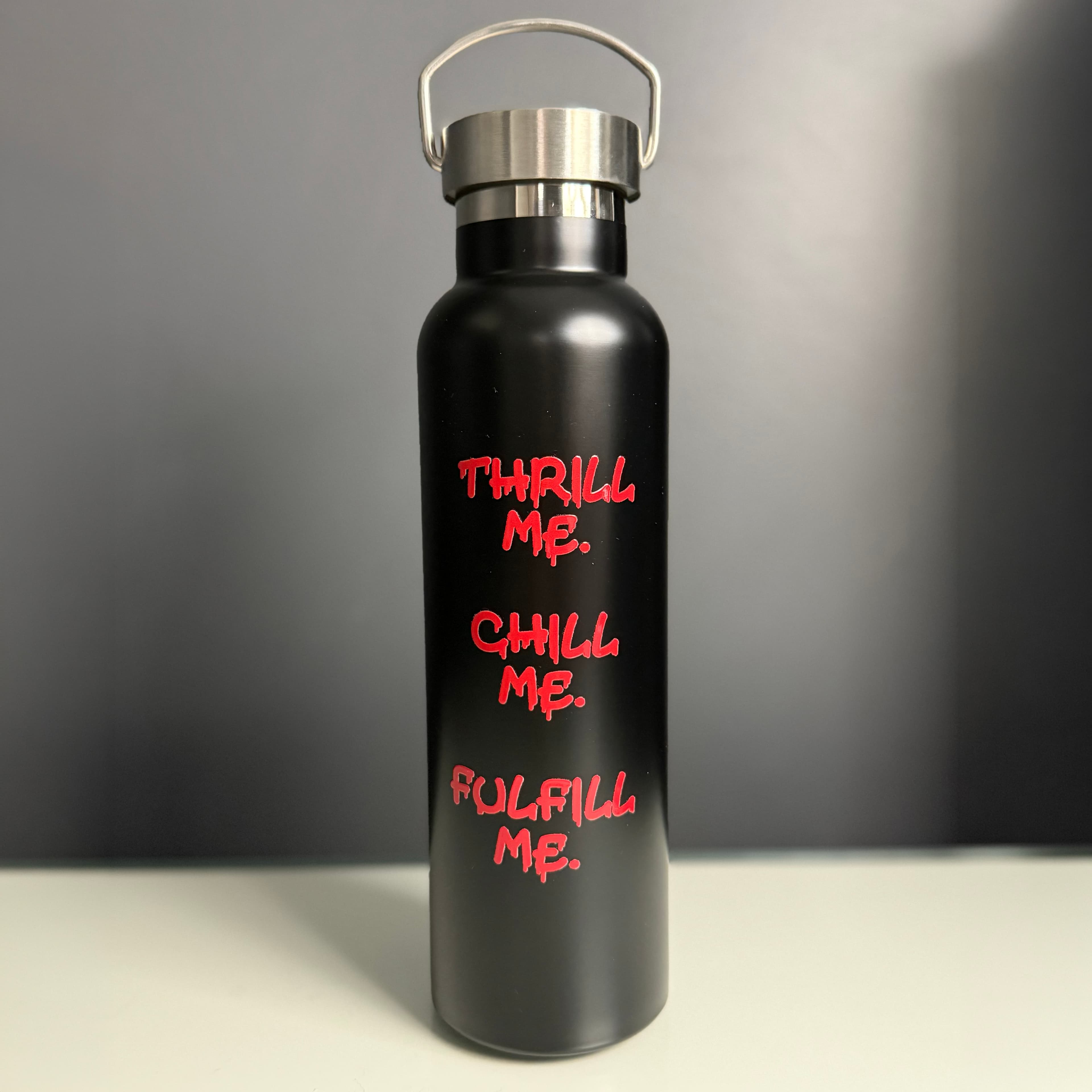 Black aluminium water bottle with a silver screw-top lid and carry handle, featuring red lettering that reads “Thrill me. Chill me. Fulfill me.”